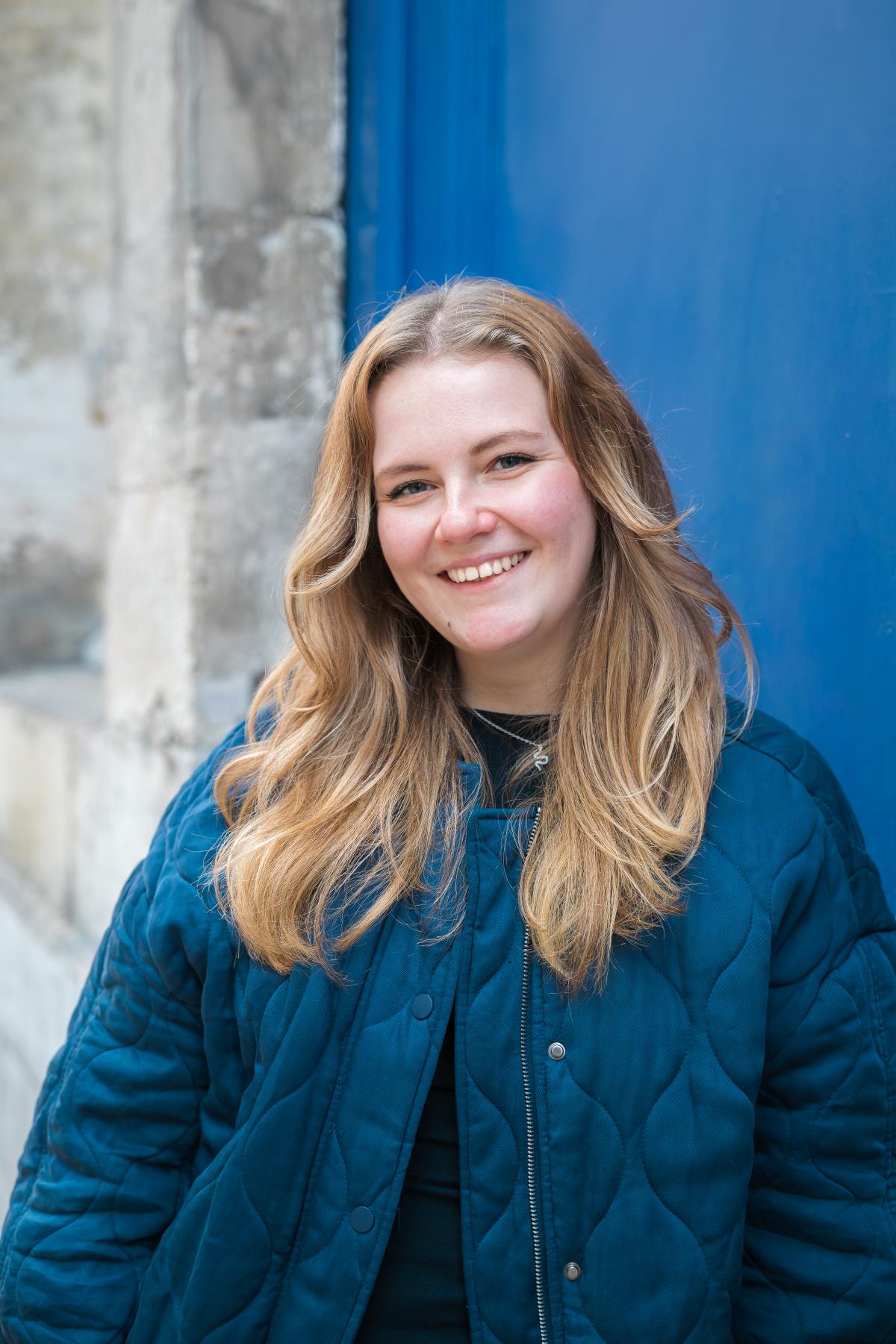 Portrait photographique d’une jeune femme blonde souriante, appuyée contre une porte bleue, portant une veste bleue