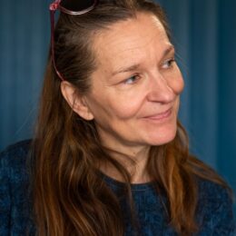 Portrait d’une femme de 50 ans vue de profil, regardant quelqu’un, cheveux bruns, portant une robe bleue, légèrement souriante, avec des lunettes sur la tête